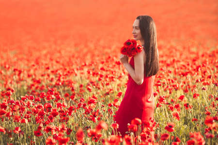 Young brunette woman 25-30 years old with long groomed hair in red dress enjoy a poppy flowers in fieldの写真素材