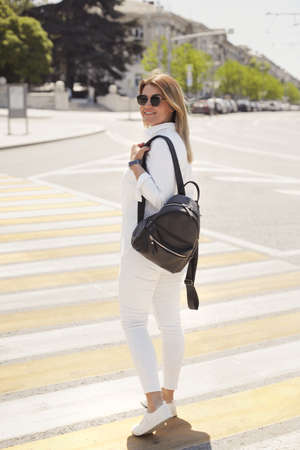 Urban portrait of happy young blonde woman 30-35 years old in white casual clothes, sunglasses and a black leather backpack walks around the city, smiling and crosses the road at a pedestrian crossingの写真素材