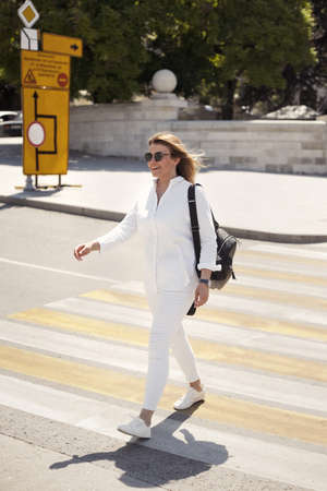 Urban portrait of happy young blonde woman 30-35 years old in white casual clothes, sunglasses and a black leather backpack walks around the city, smiling and crosses the road at a pedestrian crossingの写真素材