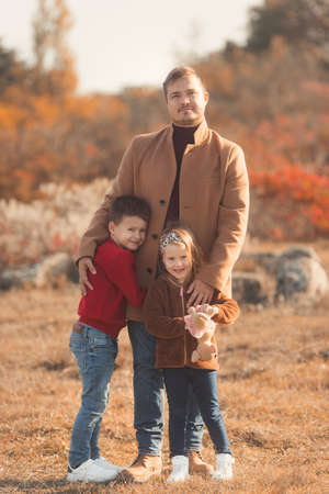 Portrait of a caucasian father hugging his children in beautiful outfits on a sunny autumn day in nature park. Family happiness concept.の写真素材