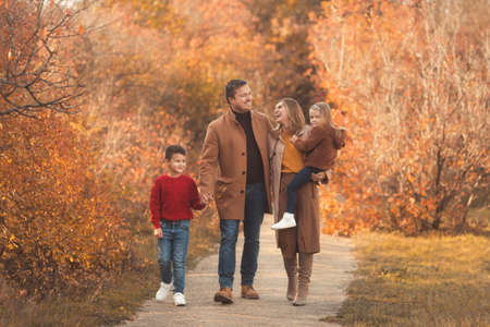 Happy family walking in the fall park. Portrait of a caucasian mother and father holding their children in beautiful outfits on a sunny autumn day in forest. Family lifestyle concept.の写真素材