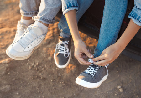 Close-up photo of legs in sneakers. The teenager boy and girl ties the laces on the sports shoes. Young people in casual blue denim clothes are sitting together outdoor.の写真素材