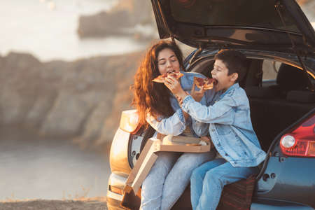 A young mother and her teenage son in denim clothes enjoy the springtime on the top of the hill, sitting in the open trunk of a car and eating pizza. Spanish tourists woman and child at vacation.の写真素材