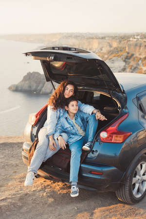 A young mother and her teenage son in denim enjoy the view of the sea and mountains on the top of the hill, sitting in the open trunk of a car and hugging together. Happy family relationship concept.の写真素材