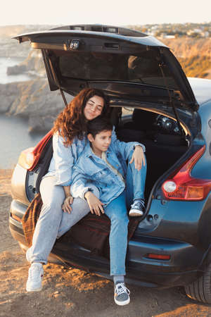 A young mother and her teenage son in denim enjoy the view of the sea and mountains on the top of the hill, sitting in the open trunk of a car and hugging together. Happy family relationship concept.の写真素材