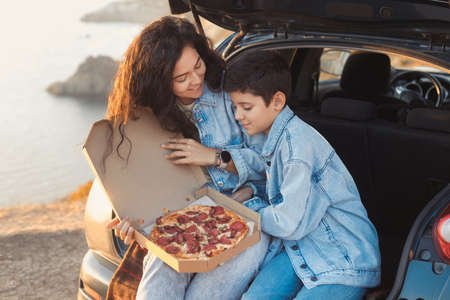 A young mother and her teenage son in denim clothes enjoy the springtime on the top of the hill, sitting in the open trunk of a car and eating pizza. Spanish tourists woman and child at vacation.の写真素材