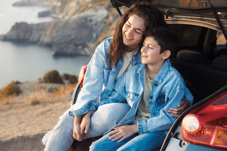 A young mother and her teenage son in denim enjoy the view of the sea and mountains on the top of the hill, sitting in the open trunk of a car and hugging together. Happy family relationship concept.の写真素材