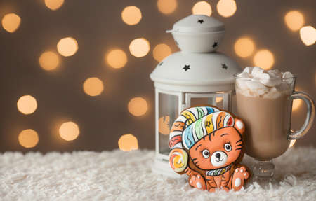 Cozy winter holiday background with gingerbread cookies tiger - symbol of new year 2022 on table, lamp with candle and cup of coffee with marshmallows above the colorful bokeh lights. Copy space.の写真素材