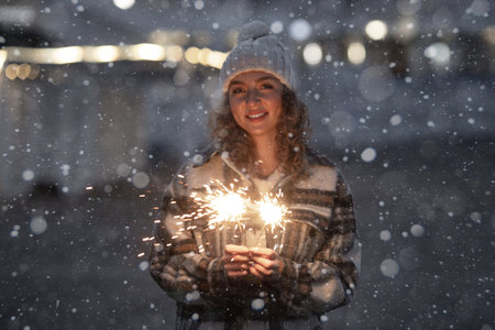 Night portrait of beautiful smiling caucasian woman 25-30 years old in fashion coat and knitted hat celebrating new year party at street with sparklers bengal lights on winter holidays outdoorの写真素材