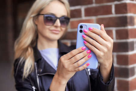 Close up photo of blonde stylish woman make photo on mobile phone. Female hands with bright neon pink manicure hold a smartphone in a purple caseの写真素材