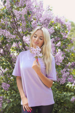 Vertical portrait of a young millennial woman 25-30 years old with closed eyes smell the aroma of flowers, blonde with long hair in a purple T-shirt in the bushes of blooming lilacs on a spring dayの写真素材
