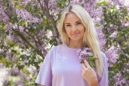 Portrait of a young woman 25-30 years old blonde with long hair in a purple T-shirt in the bushes of blooming lilacs on a spring dayの写真素材