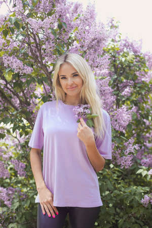 Vertical portrait of a young caucasian woman 25-30 years old blonde with long hair in a purple T-shirt in the bushes of blooming lilacs on a spring dayの写真素材
