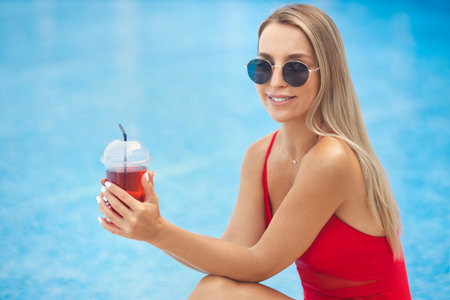 Close up portrait of young beautiful smiling girl by the pool in a red swimsuit and sunglasses, drinking detox coctailの写真素材