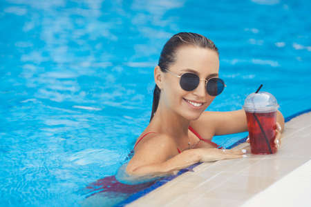 A young beautiful smiling girl is resting by the pool in a red swimsuit and sunglasses, drinking detox coctailの写真素材