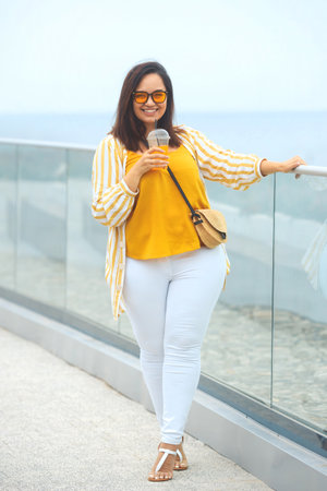 Full body portrait of happy millennial plus size woman 30-35 years old on vacation. Smiling overweight female in in yellow and white fashion clothes and sunglasses drinks juicy coctail in summer dayの写真素材