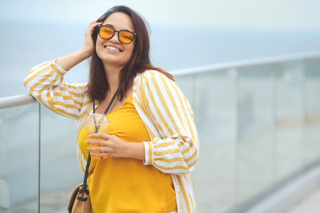 Portrait of happy positive millennial plus size woman 30-35 years old on vacation. Smiling overweight female in in yellow and white fashion clothes and sunglasses drinks juicy coctail in summer dayの写真素材