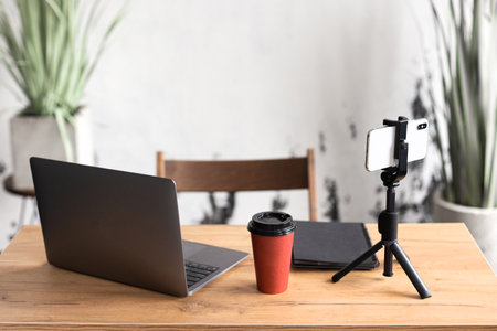 Comfortable workspace for office manager, business or freelancer in a stylish room with green plants for remote work on a laptop and online video conference using a mobile phone on a tripodの写真素材