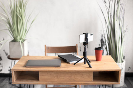 Comfortable workspace for office manager, business or freelancer in a stylish room with green plants for remote work on a laptop and online video conference using a mobile phone on a tripodの写真素材