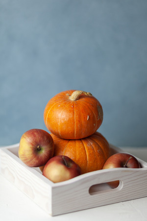 In a wooden white box are two orange pumpkins, apples. On white blue background. Autumn still-life. Halloween. Harvest. Postcardの写真素材