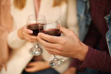 Two glasses of red wine close-up in hands outside the house. Man and woman in love make the clink of glasses. Celebrate Valentines Day, date. Warm, blurred background, countryside relaxation.の写真素材