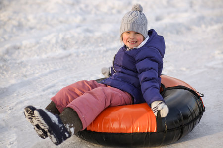 happy smiling little girl sits on an inflatable tubing. Winter fun, fun, outdoor games skating. Walk sports. Close-up rear view. Winter snow background.の写真素材