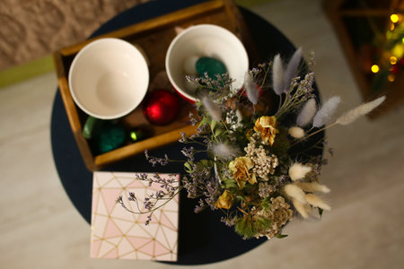 Cup and glass with print snowman on wooden table, shimmering lights. dried flowers on a wooden tray with a gift box for christmas or new year decoration bouquet compositionの写真素材