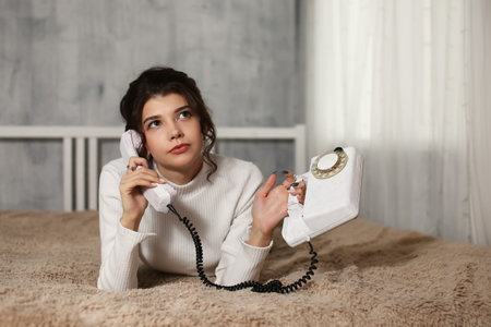 Young happy smiling woman is lying on the bed and talking on the phone. Corded vintage telephone.の写真素材