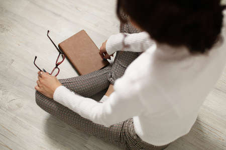 Young woman student teenager holding eyeglasses in her hands, sitting on the floor. Going to read a book, poor eyesight. The need to use glasses. View from above. Brown book, light white floor.の写真素材