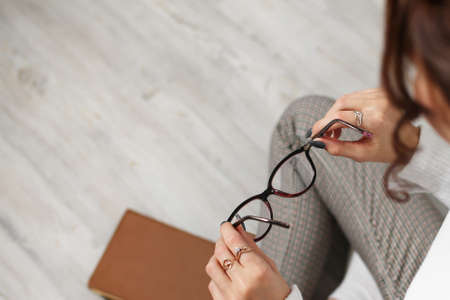 Young woman student teenager holding eyeglasses in her hands, sitting on the floor. Going to read a book, poor eyesight. The need to use glasses. View from above. Brown book, light white floor.の写真素材