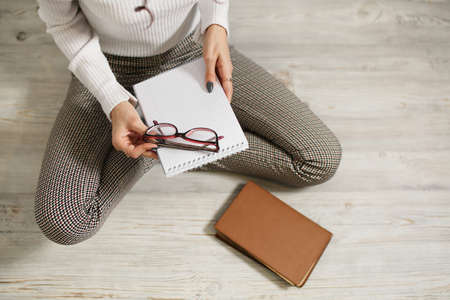 Young woman student teenager holding eyeglasses in her hands, sitting on the floor. Going to read a book, poor eyesight. The need to use glasses. View from above. Brown book, light white floor.の写真素材
