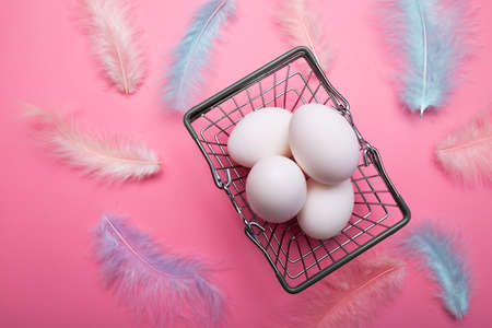 White Easter eggs in a grocery basket, shopping cart, multicolored delicate bird feathers on a pink backgroundの写真素材