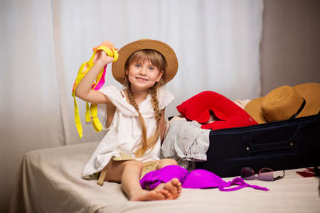 Child tourist sitting on the bed next to a suitcase, wanting to fly on an airplane, Dreaming about travel. Prepares your travel luggage bag for your adventure.の写真素材