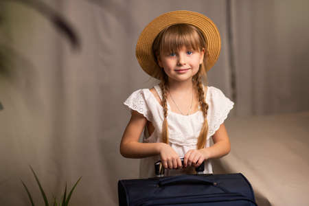 Smiling, happy girl child in a hat and a suitcase with luggage, a travel bag in her hands stands in a hotel room. Check in at the hotel, travel and trip to the sea, restの写真素材