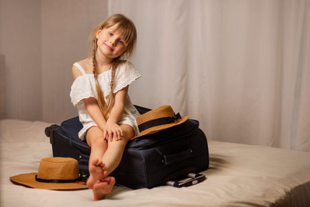Smiling, happy girl sitting on a suitcase of luggage, travel bag in a hotel room. Accommodation at the hotel, transfer and trip to the sea, rest. Straw hats lie nearby.の写真素材