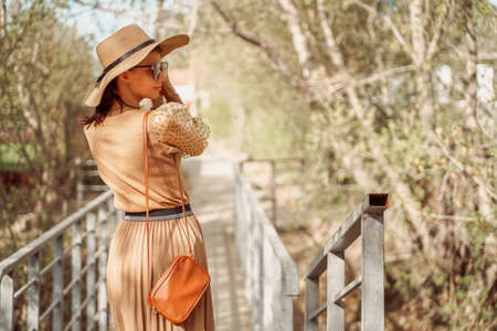 A stylishly dressed girl in a romantic beige dress with a pleated skirt and a straw hat walks along the bridge.の写真素材