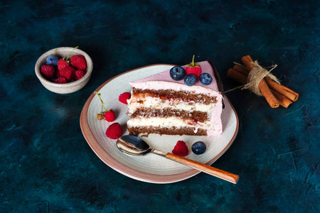 Piece of cake, chocolate biscuit with cream cheese filling on dark blue background. Berries on pink...