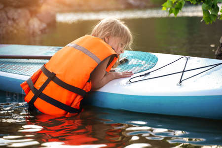 European child girl in a life jacket climbs on a SUP board floating on the lakeの写真素材
