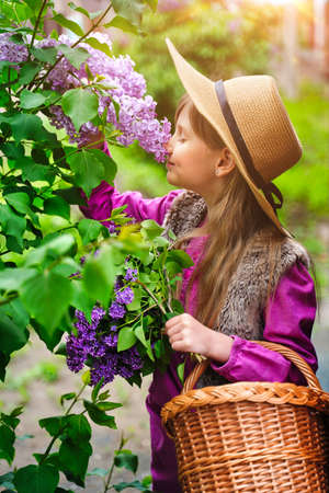Child girl in straw hat in lilac Garden. Girl smells the flowers, inhale the aroma in springtime. Gardening.の写真素材
