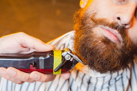 Barber cuts a beard with a trimmer to a young handsome guy with a mustache. Haircut of a beard in a...