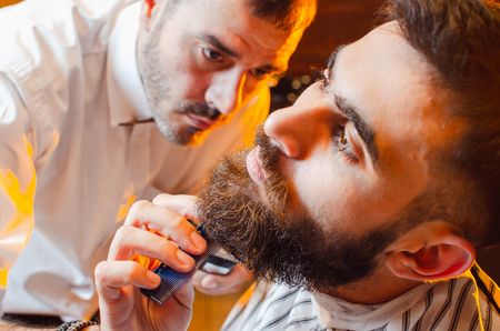 Barber cuts a beard with a trimmer to a young handsome guy with a mustache. Haircut of a beard in a male barber shop.の写真素材