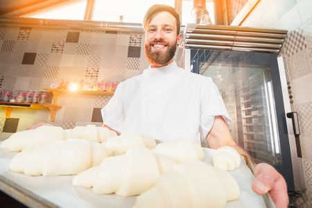 A young handsome baker is holding raw croissants of white dough in the hands of an oven. Semi-finished products from dough.の写真素材