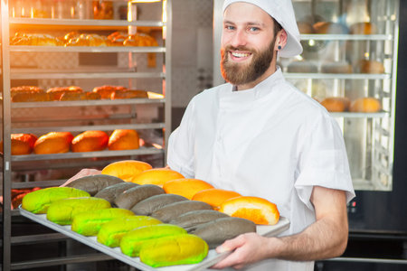 A young handsome baker is holding colorful fresh baked goods on a sheet on the background of the...