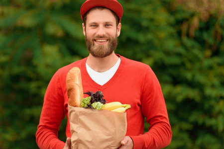 Portrait of a food delivery man in a red uniform holding a paper eco bag with groceries in his...
