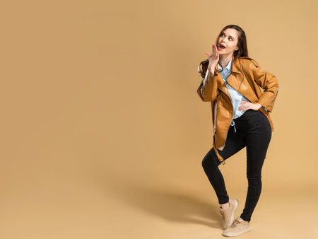 Young beautiful girl with long curly hair in a brown leather jacket covers her mouth with her hand...