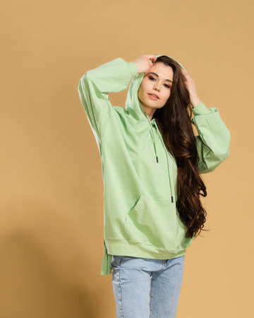 Young beautiful girl with long hair in a pistachio-colored hoodie straightens the hood on a pastel...