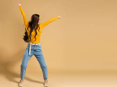 Young attractive girl with long curly hair in an orange sweatshirt is dancing with her hands raised up on a pastel orange studio background.の写真素材