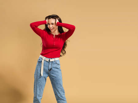 A young girl with long curly hair in a red sweatshirt on a pastel orange studio background holds her head with her hands.の写真素材