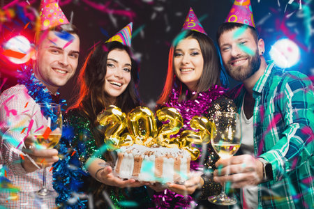 A group of young people in birthday caps with a cake in their hands with the numbers 2022 are looking at the camera against a dark background with light and musicの写真素材