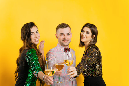Young adult girls and a guy clink glasses with white wine or champagne and look at the camera on a yellow isolated background. party of friends. Three young people with glasses in their handsの写真素材